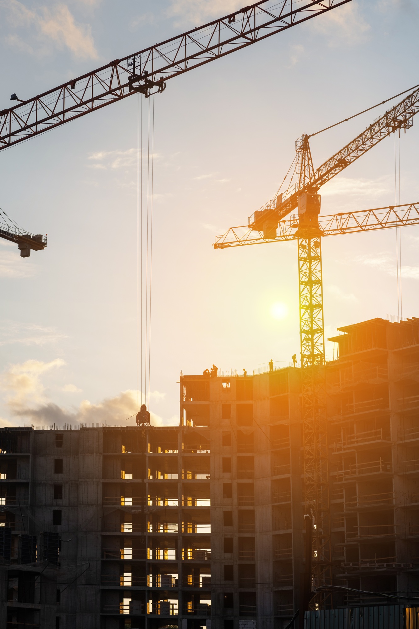 Cranes operate at a construction site during sunset, with workers actively engaged in construction